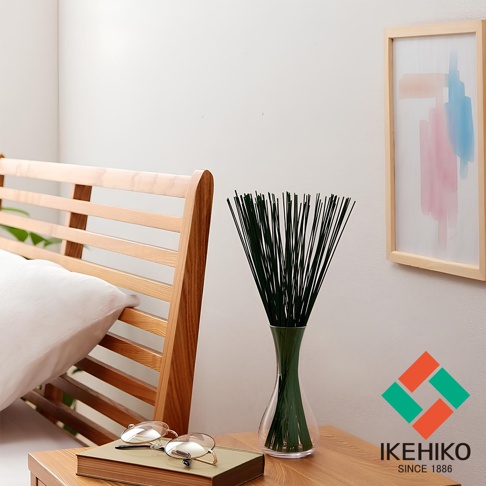 一束深綠色藺草風水清淨棒插在透明玻璃花瓶中，置於木製桌上，旁邊有書籍和眼鏡，背景是木製床架和裝飾畫。