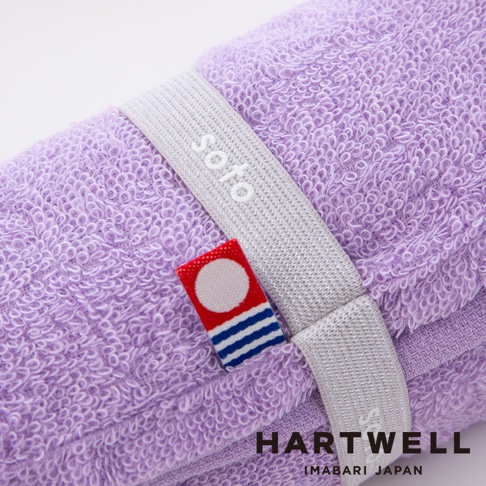 一條捲起來的淺紫色毛巾，上面繫著一條銀灰色織帶，織帶上印有 soto 字樣，織帶旁有一個紅底白圓點搭配藍白條紋的小標籤，毛巾的材質為柔軟的毛圈。