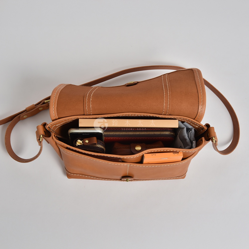 職人手作復古皮革郵差包，包身為溫暖的淺棕色皮革，縫線清晰。包內可見手機、皮夾、書本及其他小物，展現其收納空間。