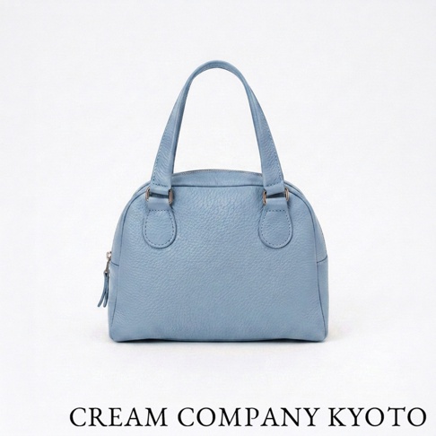 這是一個採用淺藍色荔枝紋牛皮革製作的 CREAM COMPANY KYOTO 半月型迷你手提包，包身正面有兩個對稱的提把銜接處，整體外觀簡約俐落，呈現出精緻的職人手作質感。