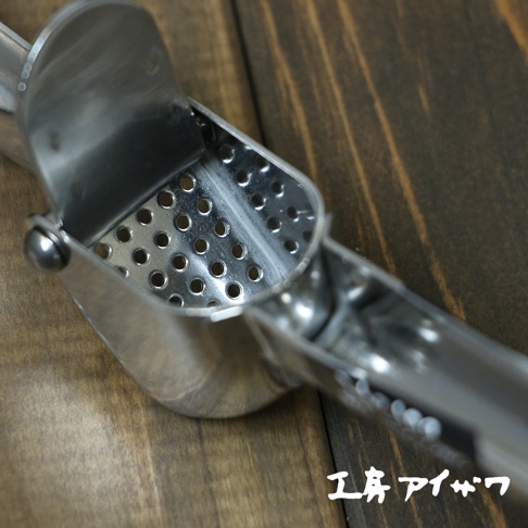 不鏽鋼壓蒜器，銀色，帶有圓形孔洞的壓榨槽，置於木紋背景上。