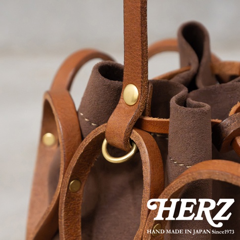 HERZ 職人手作皮革水桶包 G-98 的特寫，展示了細緻的皮革拼接、黃銅铆釘和金屬環細節。