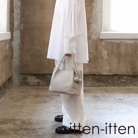 itten-itten 淺色皮革托特包，包身帶有兩個繩結裝飾，由穿著白色服飾的模特兒於室內手提展示。