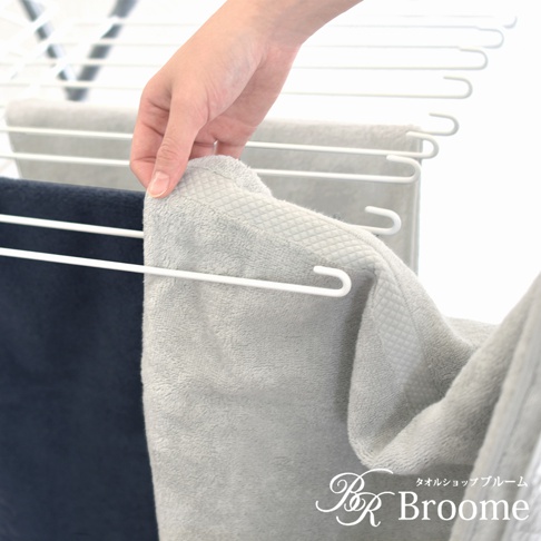 一隻手正將淺灰色、帶菱紋質感的純棉長毛巾掛在白色晾衣架上，旁邊還有一條深藍色長毛巾。