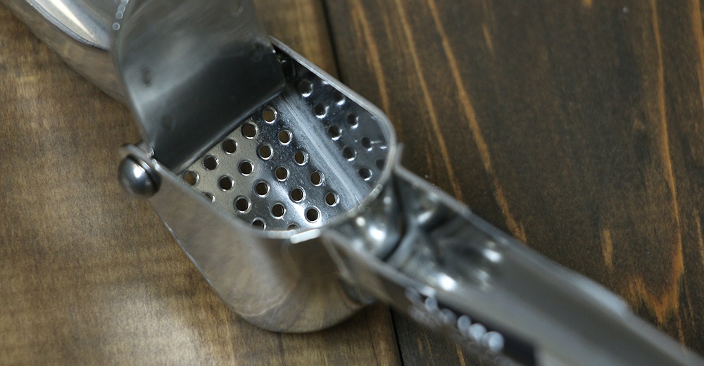 銀色不鏽鋼壓蒜器，帶有圓形孔洞的擠壓腔，置於深棕色木紋背景上。