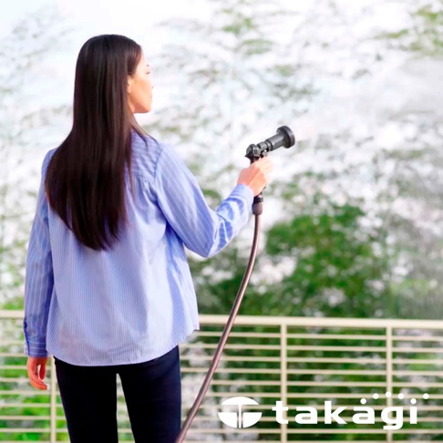 一位女性身穿藍色條紋襯衫，手持黑色灑水槍，連接一條深棕色水管，背景為模糊的綠色樹葉與白色天空。