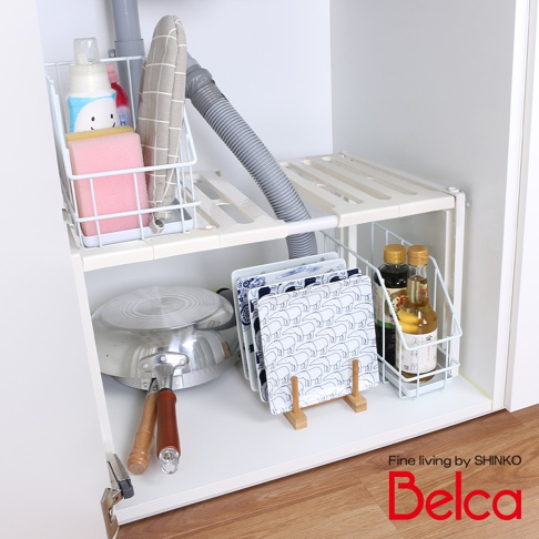 白色伸縮槽下單層站立收納架，架上放置著粉色海綿、清潔劑、灰色隔熱墊、灰色波紋軟管、兩個醬料瓶，以及一套藍白色圖案的盤子，下方還有鍋具和木質支架。