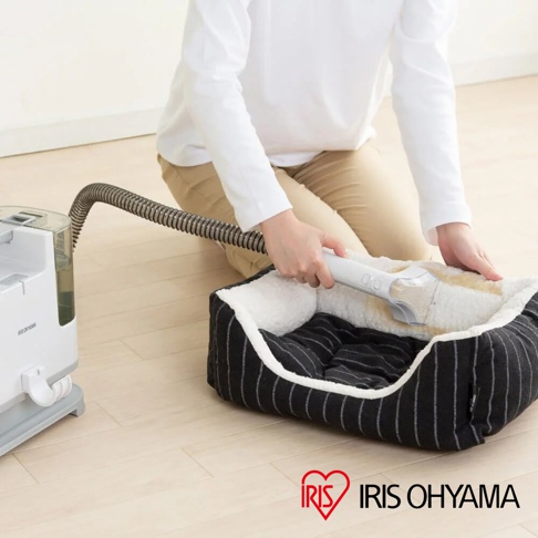 白色自動給水織物清潔機連接灰色波紋軟管，正在清潔一個帶有黑色細條紋和白色毛絨邊緣的寵物床。