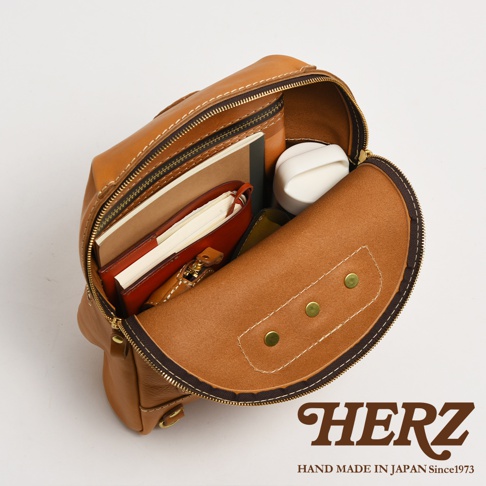 職人手作皮革大開口圓角後背包，以棕褐色皮革製成，內部裝有筆記本、皮夾和白色小物。包款設計有拉鍊開合，呈現圓潤的造型，並以白色縫線勾勒出細節。