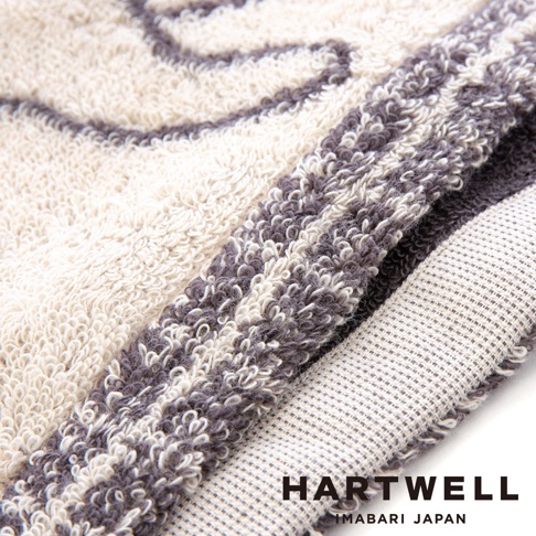 這是一張特寫鏡頭，展示了 HARTWELL 今治午睡貓咪雙色手帕的細節。畫面呈現柔軟的毛圈織物材質，手帕邊緣摺疊處清晰可見米白色與深灰色的條紋圖案，編織紋理細緻，底部印有 HARTWELL IMABARI JAPAN 的品牌標誌。