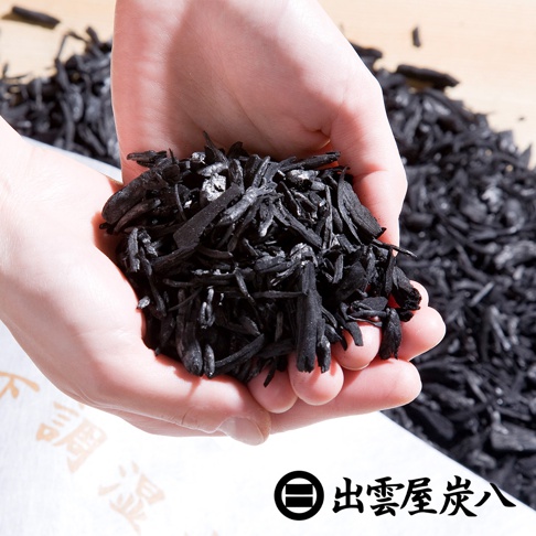 雙手捧著一把碎木炭，木炭呈不規則條狀，顏色為深黑色，部分帶有灰白色細粉。背景為模糊的木炭堆，右下角有日文品牌標誌。