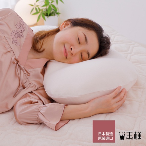 一位女性側臥於枕頭上，臉部朝向左側，閉著眼睛，面帶微笑。她身穿一件淺粉色絲綢睡衣，睡衣左肩有精緻的刺繡。枕頭為白色，材質柔軟，表面有細密的紋理。背景為鋪有白色格紋床單的床鋪，以及一盆綠色植物。