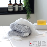 灰色沐浴巾堆疊，覆蓋豐富泡沫，產品置於白色磁磚上，背景可見三瓶深色沐浴乳與一塊黃色肥皂。