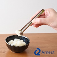 手持不鏽鋼萬用料理夾，夾取一塊白色食材，上方為白色背景，下方為木質桌面及一個黑色紋理小碗，碗內盛有白色塊狀食材。