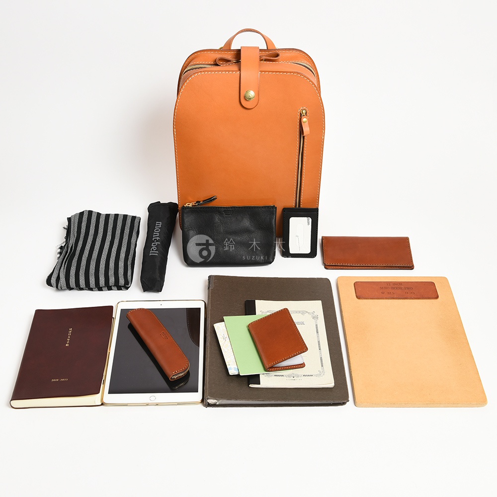 一只橘棕色皮革後背包，搭配黑色條紋圍巾、黑色摺疊傘、黑色皮革零錢包、白色證件套、棕色名片夾，以及兩本筆記本、平板電腦、棕色筆套、兩張卡片和淺棕色文件夾。