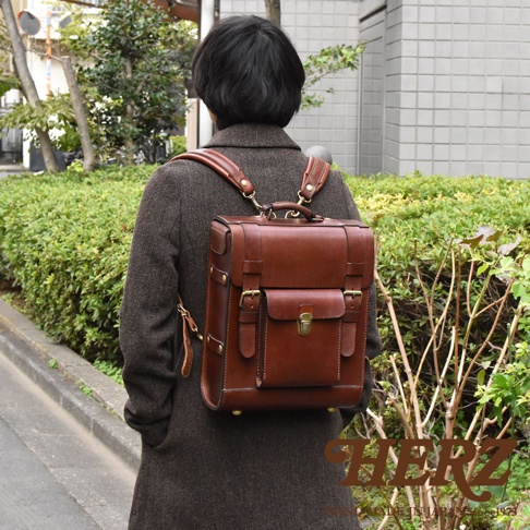 一個身穿棕色長外套的人，背著一個經典學院風的棕色皮革後背包。背包有頂部提把、兩個磁扣前袋，以及側邊的束帶和金屬扣環。