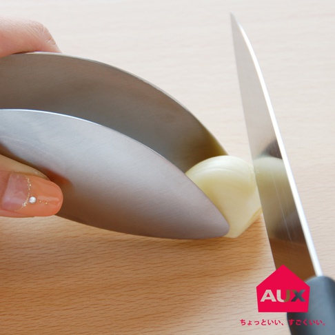 不鏽鋼調理夾夾著一顆白色蒜頭，一隻手拿著調理夾，旁邊一把刀正準備切蒜。