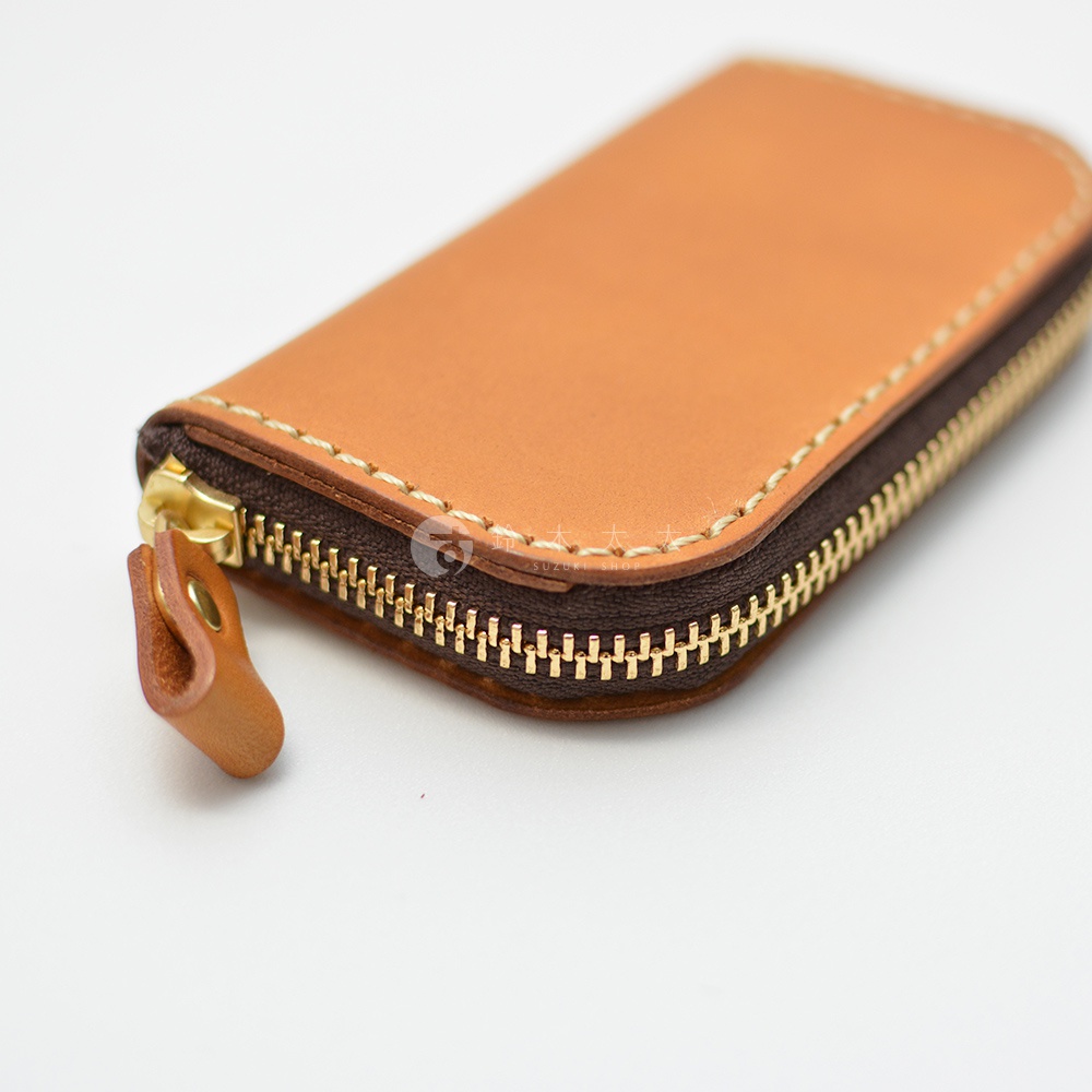 職人手作皮革橢圓形拉鍊鑰匙包，附有六個鉤子，呈現自然焦糖色，搭配金色拉鍊和縫線細節。
