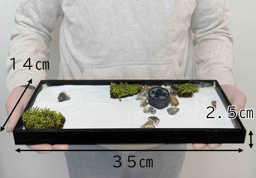 黑色木框枯山水模型，內含白色細沙，耙出漣漪紋，點綴著幾塊灰色岩石、綠色苔蘚裝飾，以及一個帶有漢字的黑色圓柱形裝飾物。