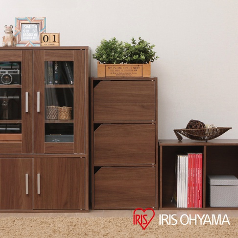 展示一個木質三層櫃，顏色為深棕色，帶有銀色把手，櫃門為透明玻璃。旁邊有一個較矮的木質櫃子，裡面放著幾本書和一個灰色收納盒。櫃子上方擺放著裝飾品和綠色植物。