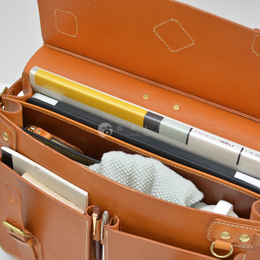 職人手作皮革旅行包 A-102，採用棕色皮革製作，包內裝有筆記本、文件夾、毛巾及黃色封面書籍，展現精緻的縫線與金屬扣環設計。