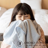 一位女子用淺藍色毛巾輕柔地觸碰臉頰，毛巾質地柔軟，帶有細緻的紋理，背景為模糊的臥室環境。