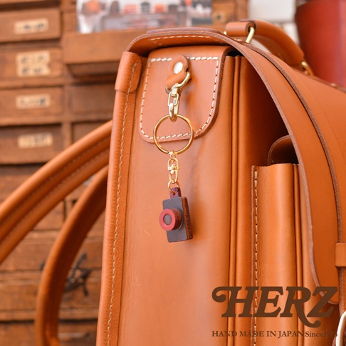 職人手作皮革相機造型鑰匙圈，以深棕色皮革製成相機主體，搭配紅色鏡頭裝飾，掛於黃褐色皮革包上，配有金色鑰匙圈及掛扣。