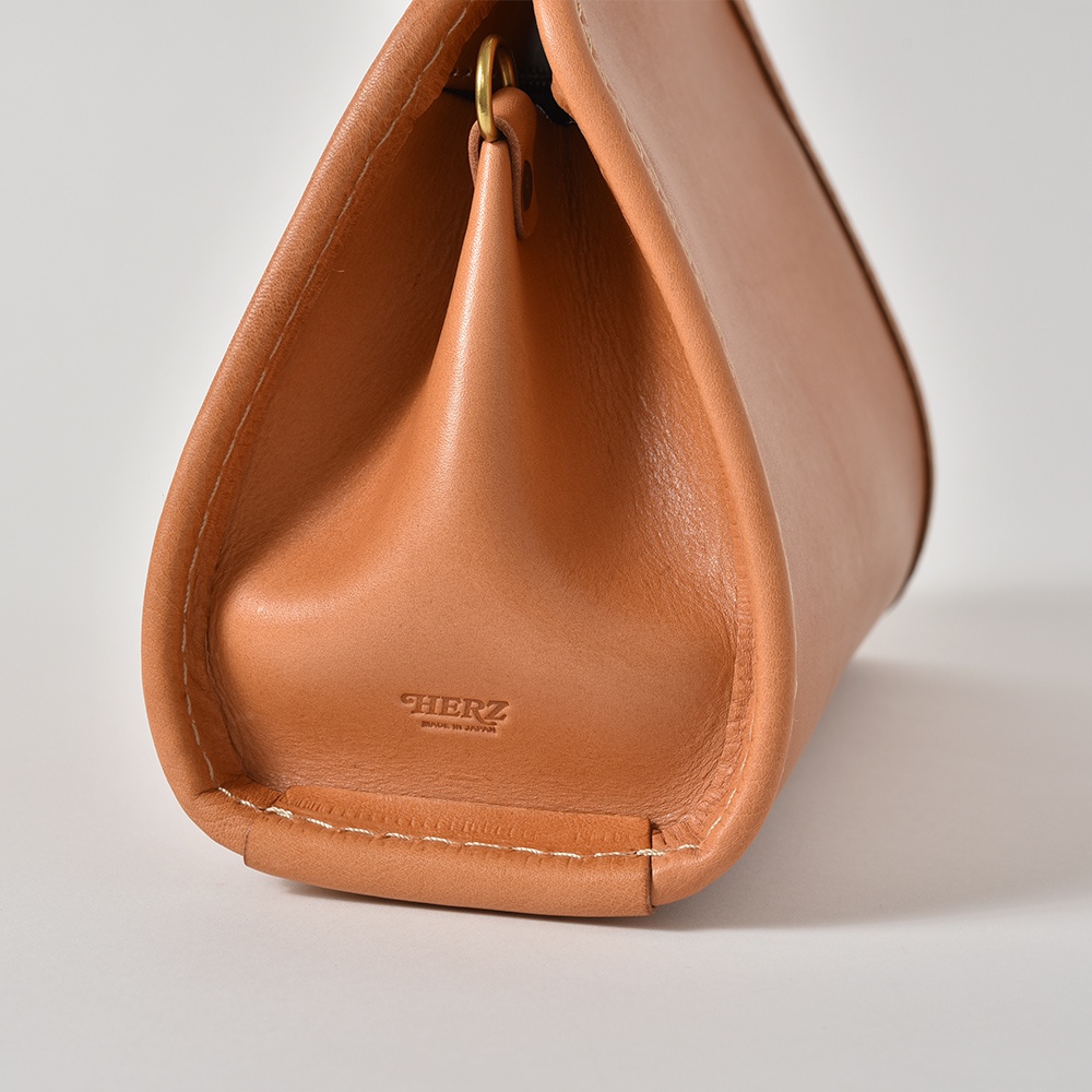 職人手作皮革單肩兩用拱型手袋，呈現溫潤的駝色調。包身以柔軟的皮革製成，縫線細膩，並帶有品牌壓印標誌，展現精湛工藝。