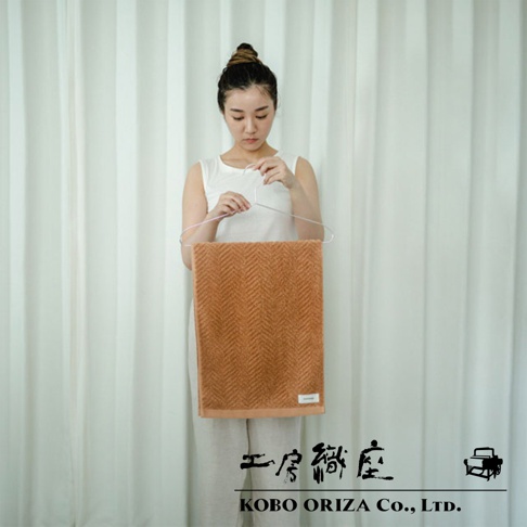 一條橘棕色人字紋有機棉浴巾，以掛架懸掛，背景為白色布簾。