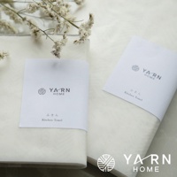 兩條白色廚房拭巾，其中一條附有印有「YARN HOME」品牌標誌與日文「ふきん Kitchen Towel」字樣的白色紙帶。背景為淺色織物，右上角有乾燥的白色小花裝飾。