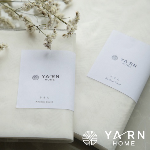 兩條白色廚房拭巾，其中一條附有印有「YARN HOME」品牌標誌與日文「ふきん Kitchen Towel」字樣的白色紙帶。背景為淺色織物，右上角有乾燥的白色小花裝飾。