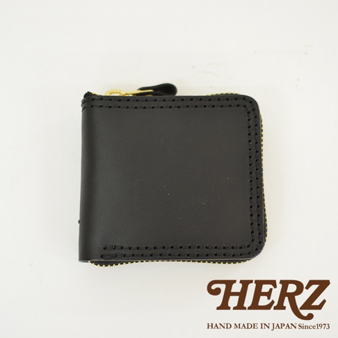 一個黑色皮革製成的HERZ職人手作全包式拉鍊短夾，採用Latigo軟皮革材質，正面可見明顯的粗縫線細節與金屬拉鍊，下方印有HERZ品牌名稱與相關文字。
