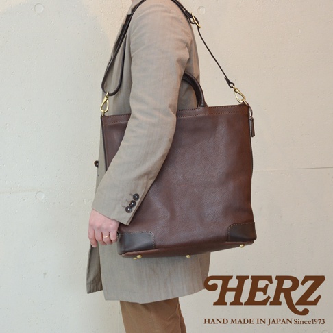 一個身穿米色外套的人側身站立，左肩斜掛著一個棕色的皮革托特包，包上有拉鍊和手提把。下方有「HERZ」的品牌標誌及「HAND MADE IN JAPAN Since1973」的文字。