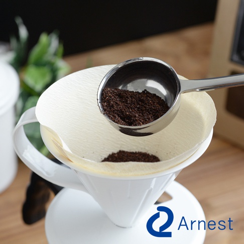 銀色不鏽鋼咖啡量匙夾，盛有細緻的咖啡粉，正準備倒入白色的濾紙杯中，濾紙杯置於白色咖啡濾杯上。