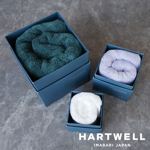 今治世紀傳承洗鍊輕盈浴巾，三款顏色：深綠、淺紫、白色，皆為柔軟材質，捲成圓筒狀，置於深藍色禮盒中，呈現精緻質感。
