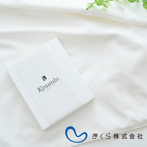 白色禮盒上印有「Kinumin」品牌標誌與「MADE IN JAPAN」字樣，置於白色絲滑織物上，背景有綠葉裝飾，底部有藍色心形圖案與日文公司名稱「まくら株式会社」。