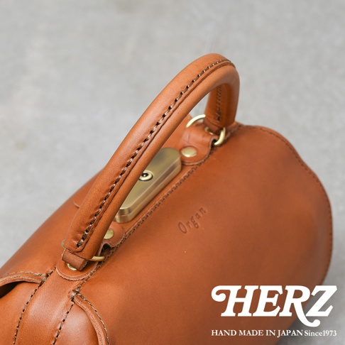 棕色皮革製成的圓筒狀手提包，採用職人手縫工藝，配有金色鎖扣和品牌壓紋。