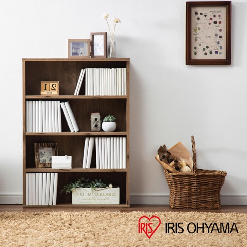 展示一個木質四層書櫃，收納多本白色封面的書籍，書櫃頂部擺放著相框和玻璃瓶，右側牆壁掛著一幅裝飾畫，地面擺放著編織籃。