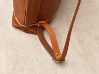 職人手作皮革後背包，呈現自然的棕褐色，皮革表面有細緻的褶皺紋理。背包肩帶為皮革材質，搭配金屬扣環，展現簡約質感。