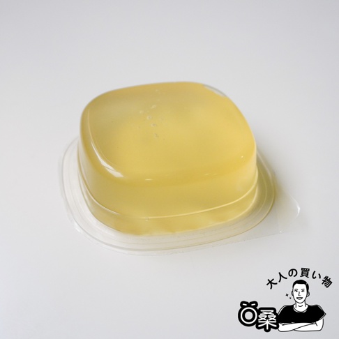 放在白色背景上的一個獨立小包裝方形葛餅，外層為透明密封塑膠殼，內含呈半透明琥珀色澤的果凍狀甜點，右下角配有大人的購物桑的黑白插圖與文字標誌。