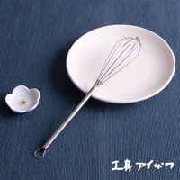 銀色不鏽鋼材質的攪拌棒，帶有圓形握柄環，置於白色圓盤上，旁邊有一個白色小碟，碟子上有花朵圖案，整體擺放在藍色紋理布墊上。