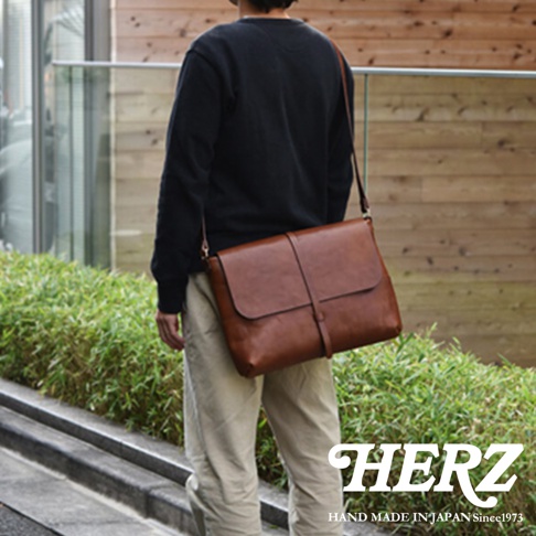 一個男人背著一個棕色皮革馬鞍包，包的正面有細長的皮帶和釦環，旁邊有綠色植物，包的右下角有 HERZ 的 Logo 和 HAND MADE IN JAPAN Since1973 的文字。