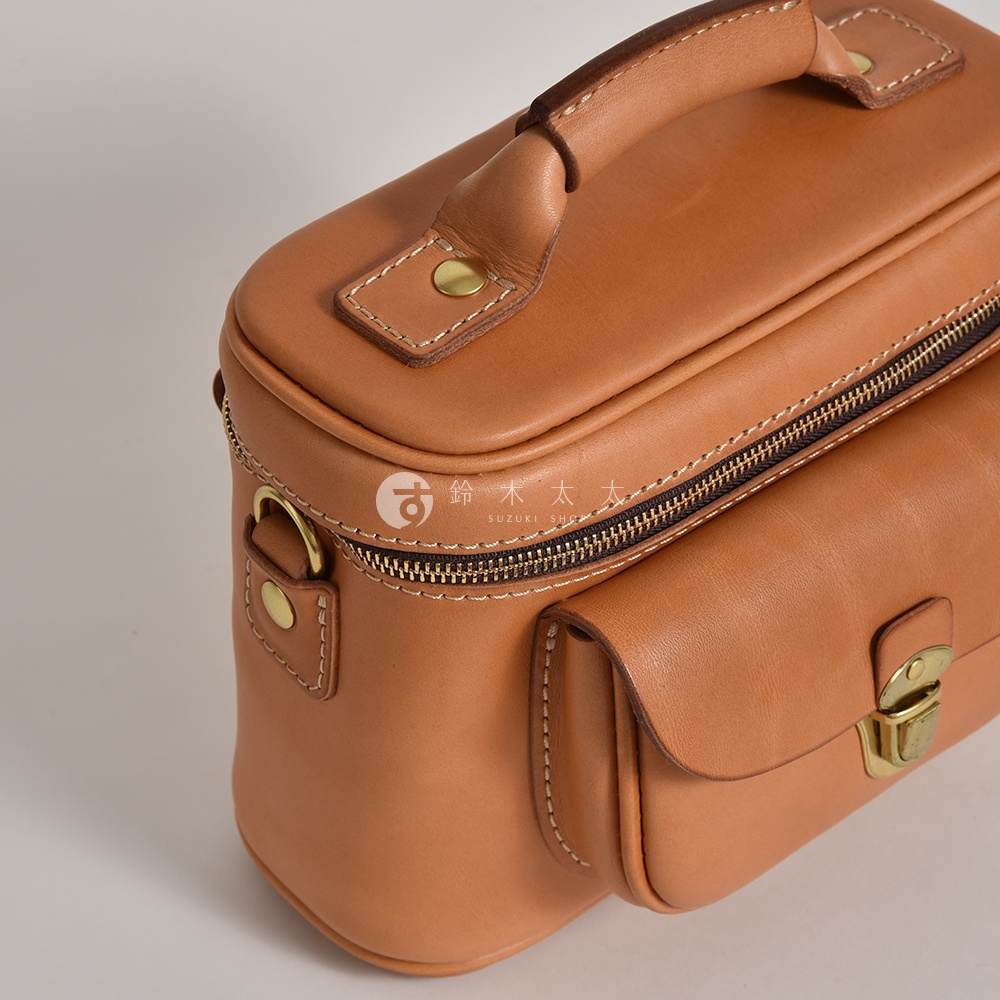 咖啡色軟皮相機包，有頂部提把、拉鍊開合，前方有磁釦翻蓋小袋，側邊有 D 型環和鉚釘細節。