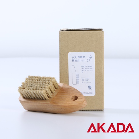 天然豬毛製成的塊莖類蔬果刷，木質刷柄呈圓潤的弧形，頂部附有密集的淺棕色豬鬃毛。一旁是印有產品資訊的牛皮紙盒。