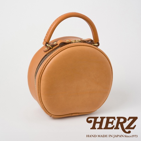 HERZ 品牌標誌，下方有 HAND MADE IN JAPAN Since1973 字樣，主體為一個褐色的皮革圓形包，帶有提把與拉鍊，風格簡約。