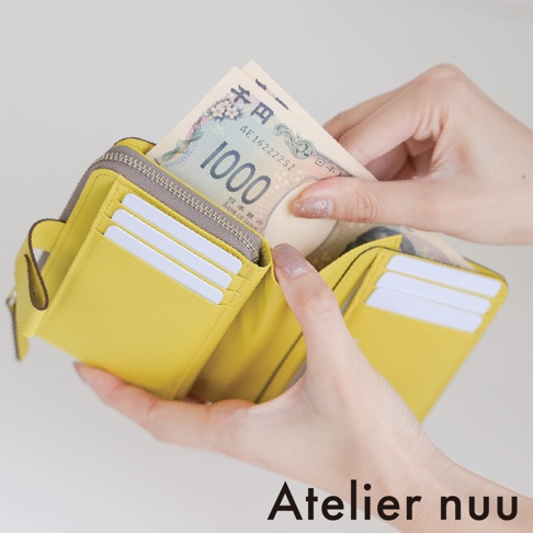 一名女性雙手展示 Atelier nuu 亮黃色皮革短夾，皮夾內裝有日幣千元鈔票，短夾兩側設有多個白色卡位，整體設計簡約精緻。