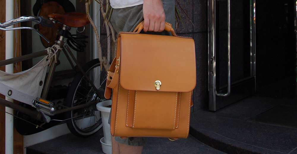 手持式棕褐色皮革後背包，具有金色鎖扣和白色縫線細節，背包有翻蓋設計和前置口袋。