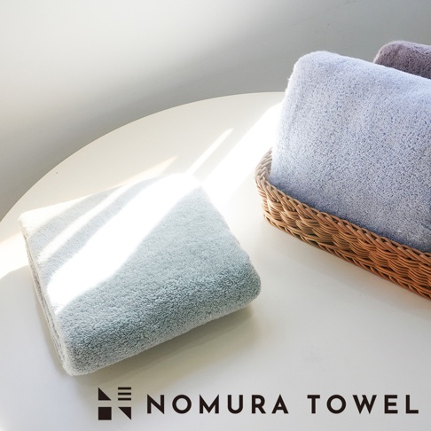 野村毛巾品牌標誌和 NOMURA TOWEL 文字，旁邊擺放著兩條疊好的毛巾，一條淺藍色，一條淺綠色，淺綠色毛巾放在白色桌面上，淺藍色毛巾放在籃子裡。