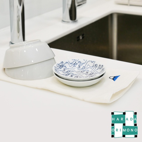 白色擦拭巾上堆疊著兩個淺白色碗，碗內放有兩個帶有藍色線條圖案的白色小盤。擦拭巾一角繡有藍色三角形圖案。