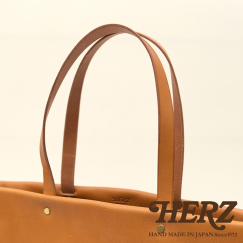 職人手作皮革極簡托特包，採用天然皮革材質，呈現溫潤的焦糖棕色。包身結構簡潔，配有兩個皮質提把，提把與包身以金屬鉚釘固定，質感樸實。